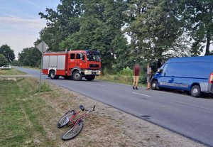 Na zdjęciu w przydrożnym rowie widnieje rower z uszkodzonym przednim kołem, po przeciwnej stronie na poboczu samochód Ford koloru niebieskiego. Na drodze pojazd Straży pożarnej