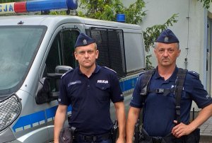 Na zdjęciu widocznych dwóch policjantów w umundurowaniu służbowym koloru niebieskiego stojącym przy oznakowanym radiowozie marki Ford