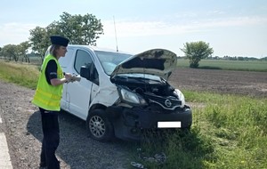 Na poboczu drogi znajduje się biały samochód osobowy marki Nissan. Posiada uszkodzenia przodu pojazdu oraz posiada zarysowania i wgniecenia prawego boku. Maska jest otwarta. Przy pojeździe znajduje się policjantka z nałożoną kamizelką odblaskową koloru żółtego.