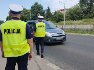 Na zdjęciu uwidoczniono dwóch funkcjonariuszy ruchu drogowego ubranych w kamizelki odblaskowe koloru żółtego. Posiadają na głowie czapki służbowe z białym otokiem. Funkcjonariusze stoją przy drodze