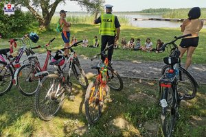 Zdjęcie poglądowe na którym uwidoczniony policjant komórki ruchu drogowego ubrany w kamizelkę odblaskową koloru żółtego na głowie posiada białą czapkę. Prowadzi prelekcje do siedzących przed nim dzieci. Na pierwszym planie widoczne rowery w tle woda oraz zakrzaczenia i drzewo