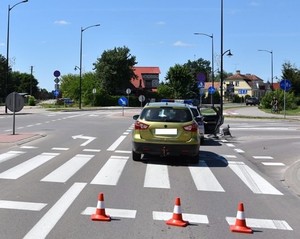 Zdjęcie poglądowe miejsca wypadku drogowego. Na zdjęciu widoczny pojazd koloru zielonego, przed nim stoi oznakowany radiowóz. Po prawej stronie leży rower.