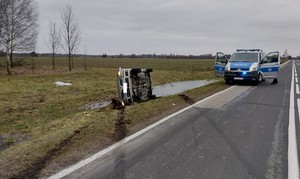 Na zdjęciu widać pojazd koloru szarego przewrócony na prawy bok. Przed pojazdem widoczne ślady hamowania. w tle radiowóz policyjny