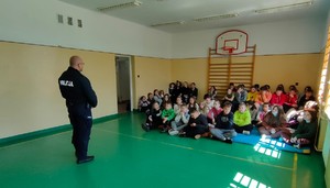 Policjant w trakcie prelekcji z uczniami. Spotkanie odbywa się w hali sportowej. Młodzież siedzi na podłodze