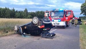 Miejsce wypadku drogowego w Hannie. Leżący na dachu ciemny samochód Audi, w tle pojazd straży pożarnej i strażacy