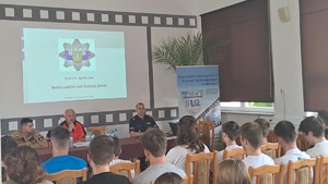 Zdjęcie z debaty społecznej we Włodawie - widok od tyłu sali młodzież uczestnicząca w debacie, trzech prelegentów - przedstawiciel Policji, Ratownictwa Wodnego i Straży Pożarnej. Za nimi ekran z gwiazdą policyjną i napisem Debata Społeczna Bezpieczeństwo nad Jeziorem Białym
