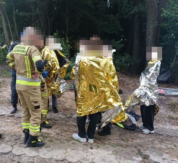 osoby poszkodowane w wyniku przewrócenia się żaglówki, stoją na plaży, obok funkcjonariusz  stoją funkcjonariusze PSP