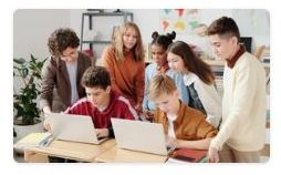 zdjęcie 2 dzieci siedzących przy laptopach, za nimi stoi piątka innych dzieci