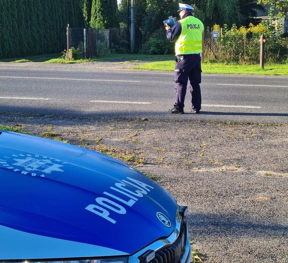 Europejski Tydzień Mobilności – akcja Prędkość - na zdjęciu fragment samochodu policyjnego i policjant prowadzący pomiar prędkości za pomocą trzymanego w ręku urządzenia