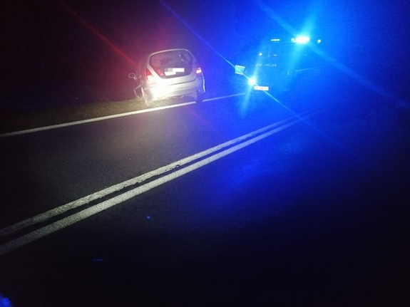 Wykonane w nocy zdjęcie z miejsca zdarzenia na którym znajduje się tył samochodu Suzuki koloru szarego przy drodze krajowej numer 82 w Kołaczach. W tle radiowóz policyjny na włączonych światłach błyskowych