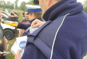 Policjant ruchu drogowego rozdający ulotki i naklejki z napisem Mistrzu Zwolnij, w tle drugi policjant bada kierującego na stan trzeźwości