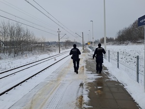 Dwóch policjantów  patroluje teren ośnieżonych peronów kolejowych. W tle słupy trakcyjne i oświetleniowe