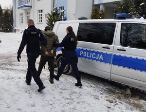 Tymczasowo aresztowany 45 - latek doprowadzany przez dwóch policjantów do zaparkowanego radiowozu